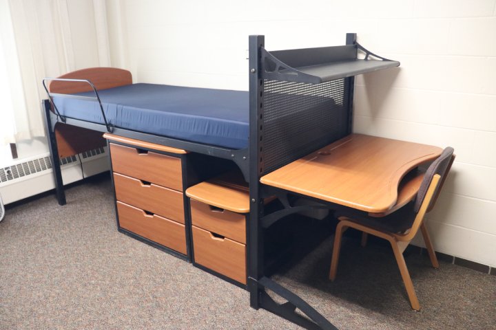 A dorm room featuring a lofted bed with a blue mattress and wooden drawers underneath. Next to the bed is a study desk with a chair, creating a compact living and study space.